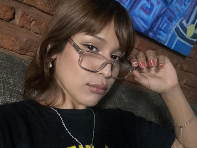Photograph of KiaraHernandezX with light brown skin, wearing glasses, black t-shirt, red nail polish, and a gold necklace, holding glasses with her right hand, brick wall and blue painting background.