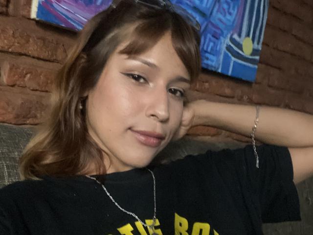 Photographie de KiaraHernandezX, avec des cheveux bruns mi-longs, portant un t-shirt noir, appuyée contre un canapé gris, une main derrière la tête, léger sourire, sexy, éclairage tamisé, fond de mur en briques, pose décontractée..