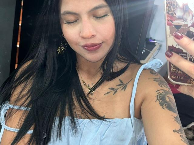 NyraVanel, con cabello negro largo, ojos cerrados, vistiendo un top blanco sin tirantes, un collar dorado y aretes de mariposa. Tatuaje visible en el hombro derecho. Se está tomando un selfie con un teléfono en la mano derecha..