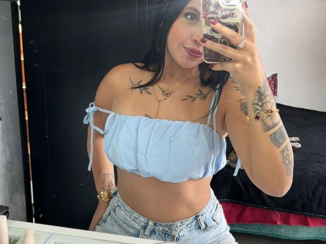 NyraVanel, sexy brunette with tattoos, takes a mirror selfie. She's wearing a white off-shoulder top, showing ample cleavage, and light blue jeans. Her hand is on her phone, lips slightly parted.