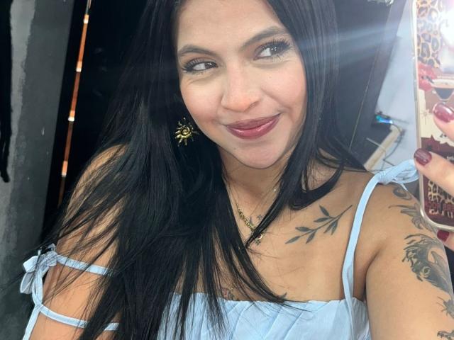 Close-up of NyraVanel, a woman with long black hair, wearing a white off-shoulder top, smiling with red lipstick. Visible tattoos on her shoulder, sunbeam on her face, holding phone.