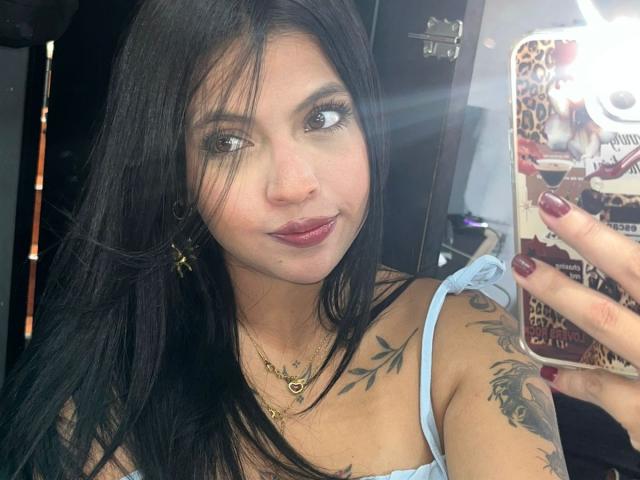 NyraVanel, avec ses longs cheveux noirs, sa peau brun clair et ses yeux foncés, porte un haut à bretelles bleu clair, un collier en or, et des tatouages sur son épaule et son bras. Prendre un selfie, ongles rouges, expression sexy.