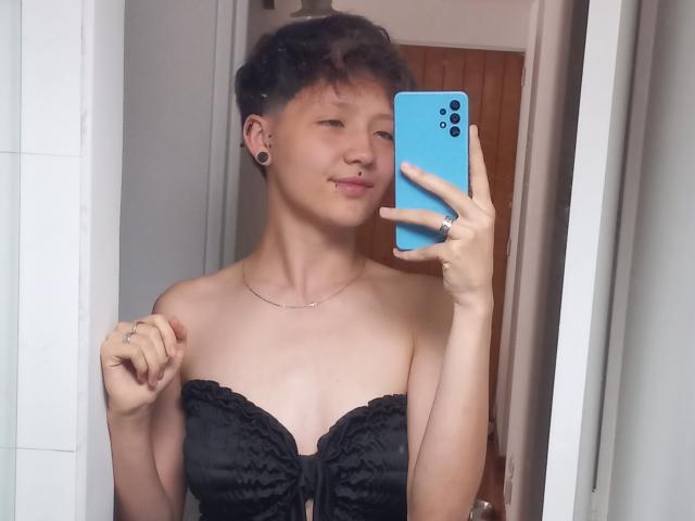 LissyLuxe se toma un selfie con un iPhone azul, vistiendo una blusa negra de encaje sin tirantes, con cabello corto y oscuro, y con aretes negros. Sosteniendo el teléfono en su mano derecha, sonriendo..