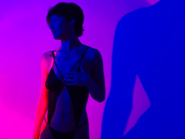 Fotografia em silhueta de LissyLuxe vestindo lingerie preta, em pé contra um fundo vibrante em rosa e azul neon. Ela está segurando o peito, com a sombra de outra figura à direita. Muito quente.