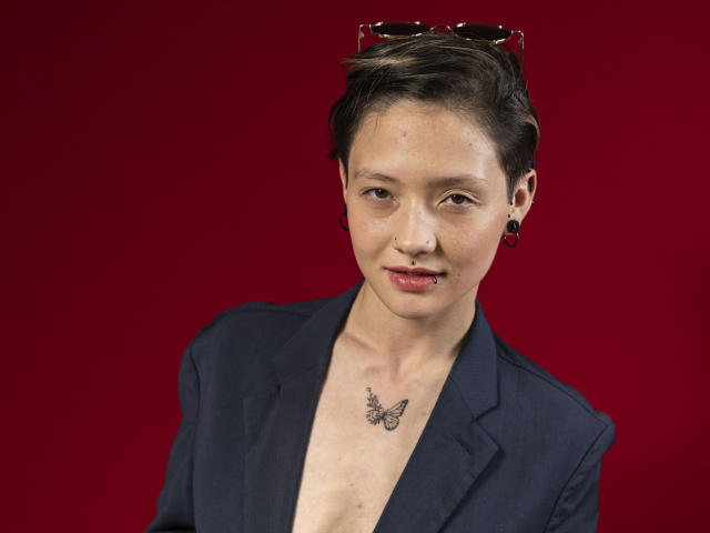 Portrait numérique de LissyLuxe, une personne mince avec des cheveux noirs courts, portant un blazer noir et sans chemise, exposant une tatouage de papillon sur leur poitrine. Fond rouge, lunettes de soleil sur le dessus.