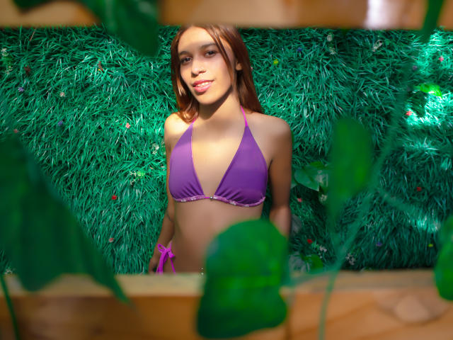 Fotografie von Valerie Woods mit heller Haut und braunen Haaren, die ein violettes Bikini-Oberteil trägt und vor einem grünen, buschigen Hintergrund steht. Wand. Sie hat einen schlanken, attraktiven Körperbau mit kleinen Brüsten. Helles Licht..