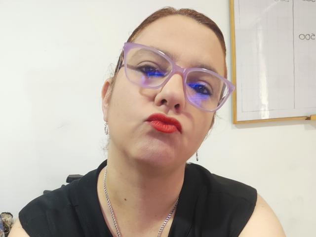 Close-up of AmeliaWilson, fair-skinned with red lipstick, wearing transparent purple glasses and a black sleeveless top. She has a subtle pout, looking slightly downward, with a silver necklace.