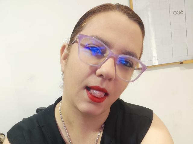 Primo piano di Amelia Wilson, una donna formosa con la pelle chiara, rossetto rosso e occhiali con montatura blu. Ha la lingua leggermente fuori, indossa una maglietta nera e una collana d'argento. Pene visibile.