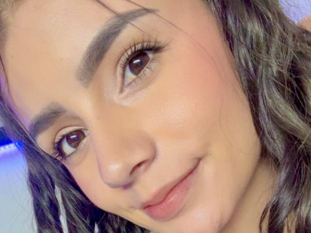 Primer plano del rostro de KimmyK, con cabello castaño ondulado, ojos marrones y una sonrisa sutil. La piel de KimmyK es suave, con un maquillaje ligero y cejas naturales. Tensión sexual implícita a través de una mirada íntima..