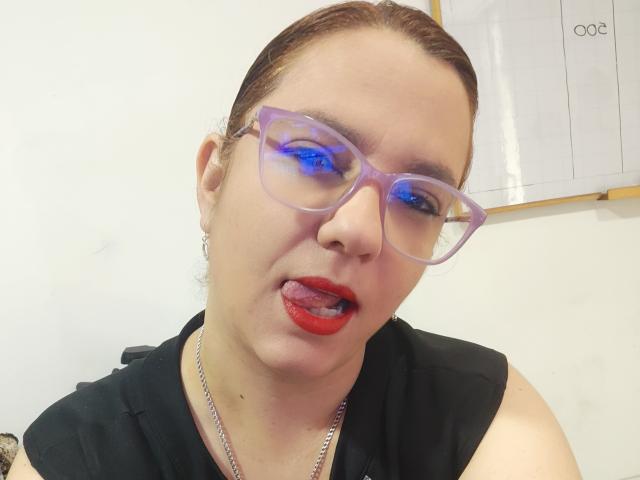 Close-up van Amelia Wilson, met een lichte huid en bruin haar, die transparante, blauw getinte brillen draagt en rode lippenstift, tong iets te groot, zwart topje zonder mouwen, witte achtergrond, sexy uitdrukking, blauwe oogreflectie..