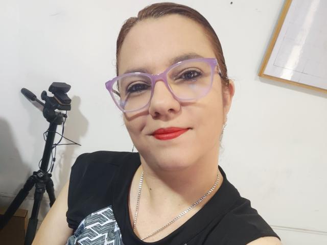 AmeliaWilson with light skin and red lipstick, wearing purple glasses and a black shirt with a white pattern, stands in front of a tripod, slightly smiling, exuding casual confidence.