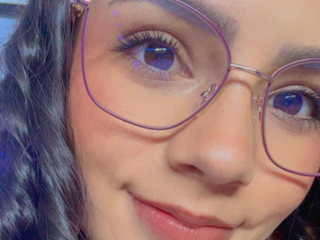 Close-up foto van KimmyK's gezicht met golvend zwart haar, die een grote bril met paarse rand draagt, blauwe ogen en een lichte glimlach. Haar huid is glad, haar lippen zijn lichtjes open, wat een subtiele seksuele spanning suggereert..