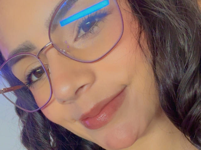 Close-up van KimmyK's gezicht, lichtbruine huid, zwart golvend haar, draagt grote ronde brillen met een blauwe reflectie, glanzend. roze lippen, lichte grijns, zichtbaar kuiltje in de wangen, zacht, natuurlijk licht.