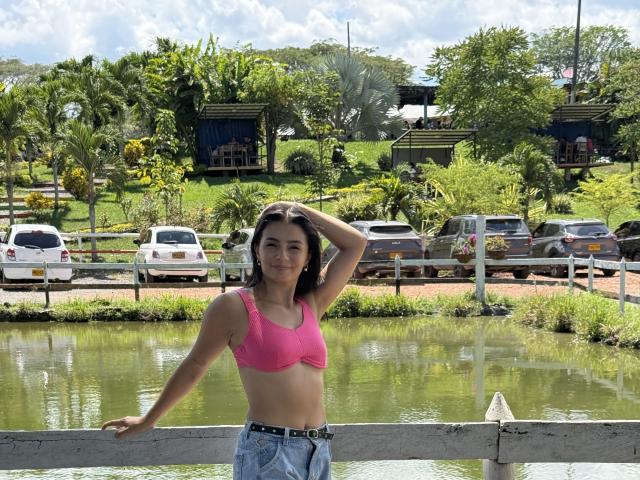 KimmyK, con cabello oscuro de longitud media, está de pie junto a un río, con la mano en el cabello, vistiendo un top deportivo rosa y jeans azules. Fondo: coches, árboles verdes, día soleado. Ligera tensión sexual..