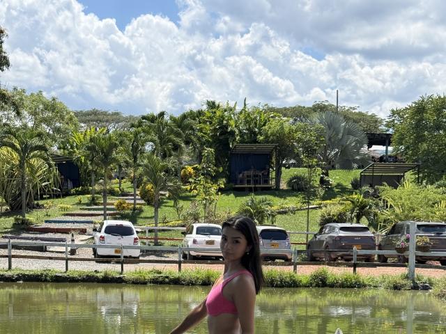 KimmyK, in piedi vicino a uno stagno, indossa un top da bikini rosa, ha lunghi capelli neri, una figura snella e sta guardando indietro. con una spalla leggermente sollevata e un'espressione un po' provocatoria. Auto e alberi sullo sfondo.