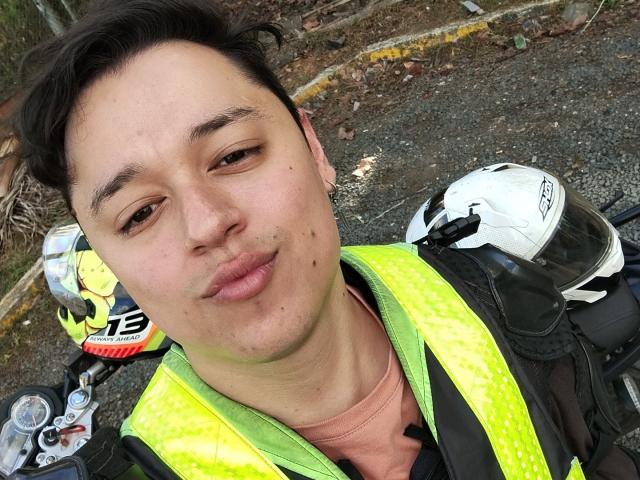 Close-up selfie van SaidHot met lichte huid, kort zwart haar, die een neongele veiligheidsvest draagt over een roze shirt. Witte helmen op de achtergrond. Buiten, een weg van grind. Een heel serieuze blik..