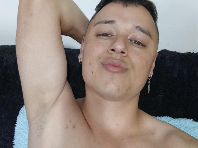 Close-up de SaidHot, um homem sem camisa, com cabelo preto curto, pele clara e pequenas manchas na face. Ele está posando com um braço atrás da cabeça, franzindo os lábios. Penísc não visível.