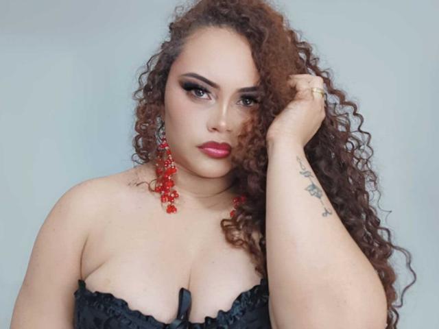 Fotografia di LunaRhose con capelli castani ricci, rossetto rosso e top in pizzo nero. Indossa degli orecchini rossi, guarda in modo seducente, con una mano tra i capelli. Pelle chiara, tatuaggi sull'avambraccio. Incredibilmente attraente..