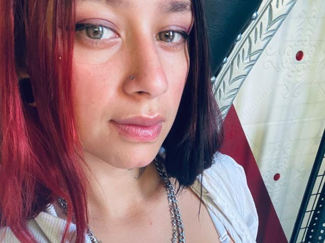 Fotografía de primer plano de RoxanaSanchezz, de piel clara, con cabello rojo y negro, un pequeño piercing en la nariz y labios rosados y brillantes. Ella mira ligeramente hacia un lado, vistiendo una blusa blanca y un collar de plata. Tensión sexual.