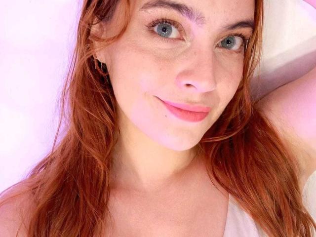 Primer plano de Geminia, una mujer de piel clara, con cabello rojo, ojos azules y pecas, sonriendo ligeramente. Su brazo derecho está levantado, mostrando sus pequeños senos y un atisbo de su pezón rosado..