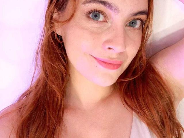 Close-up of Geminia, a young woman with fair skin, red hair, and blue eyes, smiling slightly. Her right arm is raised, showcasing her freckled face and pink lips. Fucking hot.