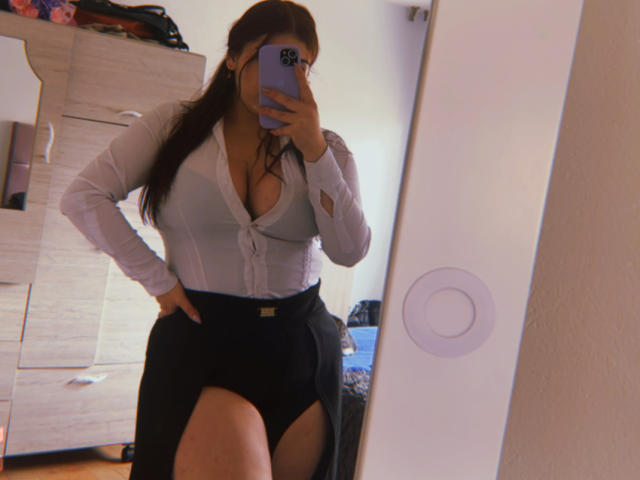 VenusxMusa se tient devant un miroir, un téléphone à la main, vêtue d'un chemisier blanc moulant avec un décolleté plongeant et d'une jupe noire. Avec une fente haute, qui dévoilait sa cuisse. Réflexion dans un miroir. Cadre d'une chambre à coucher..