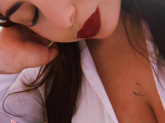 Close-up van het gezicht van VenusxMusa, met een donkerrode lipstick, halfgesloten ogen, donker haar, een wit shirt, een hand in de buurt van het oor, een halve maan. Maantatoeage op de borst. Seksuele spanning, licht verleidelijke pose, donkere make-up.