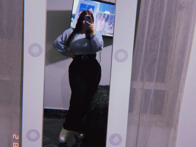 VenusxMusa se toma un selfie frente al espejo, de pie con las manos en las caderas, vistiendo una camiseta gris de manga larga y pantalones negros de cintura alta. falda, botas blancas. Se refleja una televisión en el fondo. Dedos sobre el teléfono..