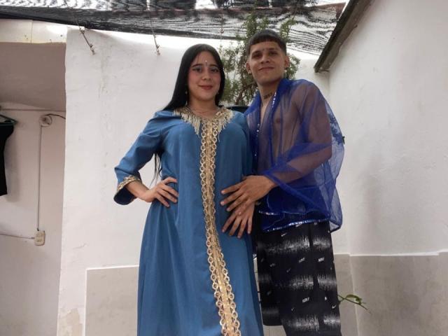 AnjaliRohan with long black hair stands in a blue salwar kameez with gold trim, hand on hip. A guy in a blue semi-sheer shirt and black pants stands close, hand on her waist.
