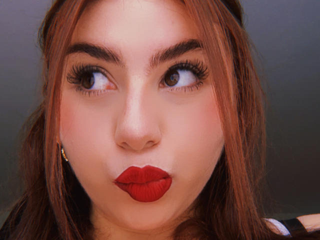 Close-up van VenusxMusa met opvallende rode lipstick, dikke zwarte eyeliner en lange wimpers. Haar bruine haar omlijst haar gezicht, en ze kijkt recht in de camera met een lichtjes fronsende uitdrukking. Lichte huid, donkere achtergrond.