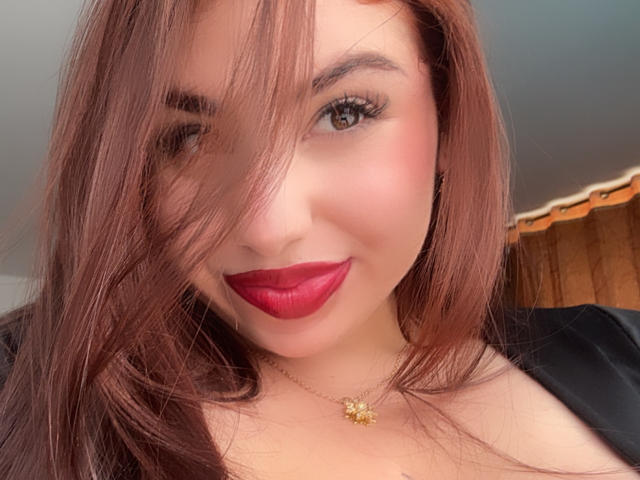 Gros plan de VenusxMusa avec du rouge à lèvres rouge, des cheveux bruns qui couvrent partiellement le visage, des yeux marrons, portant un haut noir et de l'or. Collier avec un pendentif en forme de soleil. Tension sexuelle évidente, sourire légèrement séduisant. Éclairage vif..