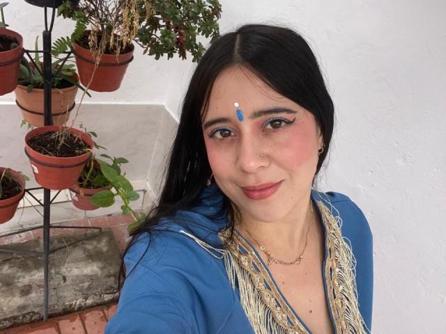 AnjaliRohan with dark hair, blue traditional outfit, gold trim, and blue bindi. Potted plants in the background. She has a subtle smile, looking directly at the camera. Fucking cute.