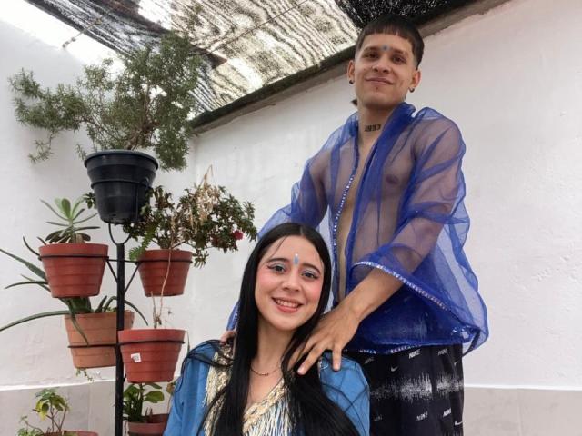 AnjaliRohan está detrás de otra AnjaliRohan, que está sonriendo y lleva una chaqueta azul. AnjaliRohan, de pie, lleva una camisa azul translúcida que revela un bulto en sus pantalones negros. Plantas en macetas en el fondo.