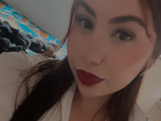 Fotografía de primer plano de VenusxMusa, una mujer con piel clara, lápiz labial rojo oscuro y cabello marrón oscuro. Ella tiene cejas pobladas y ojos oscuros. El fondo incluye una cama con un patrón azul y naranja. Increíblemente atractivo/a..