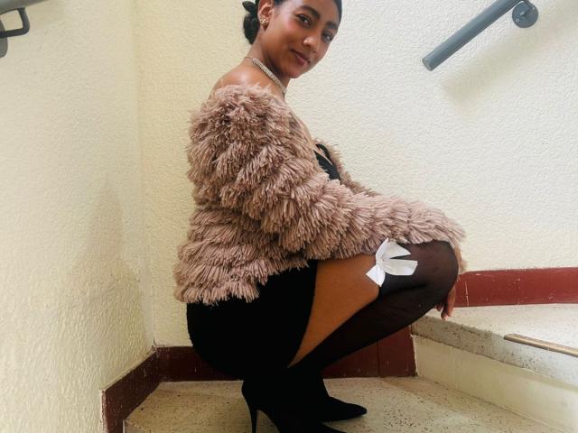 NatalyBlonder, a black woman, squats in a corner, wearing a fluffy brown jacket, black skirt, and high heels. Her black thigh-high socks have white bows. She's looking back with a seductive smile.