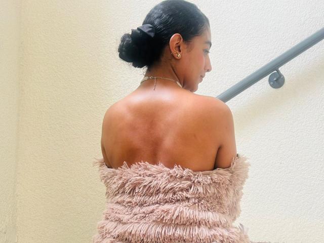 NatalyBlonder, met donkere huid en zwart haar in een knot, kijkt naar rechts en draagt een roze, pluizige top met een open schouder. Haar rug is blootgesteld, waardoor gladde huid en een sensuele pose zichtbaar zijn. Helke lichtbron..