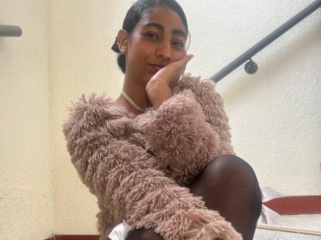 NatalyBlonder sits confidently, hand under chin, wearing a fluffy pink sweater and black tights. She has dark skin, dark hair, and a slight smile. Bright light from window behind her.