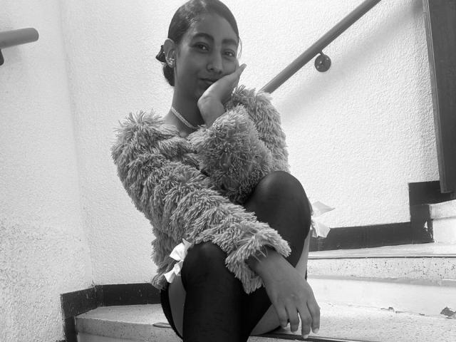 Black-and-white photo of NatalyBlonder, sitting on stairs, legs crossed, hand resting on chin, wearing a fuzzy sweater, dark pants, looking at camera with a subtle smile. Bright background light.