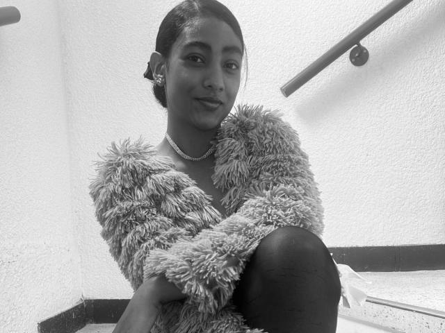 Black and white photo of NatalyBlonder with short hair, wearing a fluffy fur coat, sitting with legs crossed, subtle smile, necklace, earrings; bright light in background, slight shadow on right.