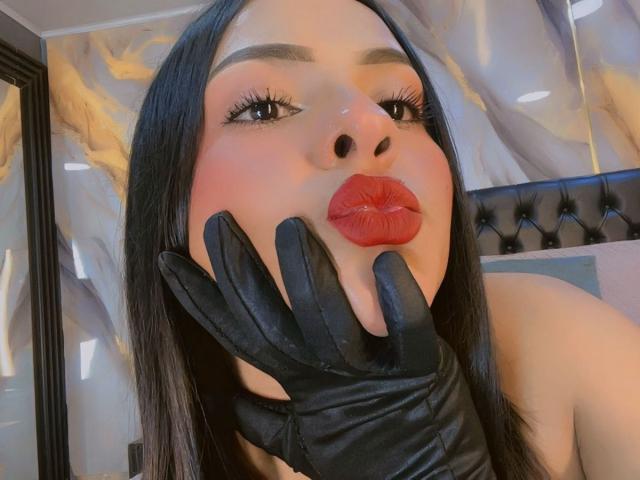 Photo en gros plan de LindaFranco, avec des cheveux noirs, portant des gants en cuir noir, appuyant son visage contre eux, avec un regard vif. des lèvres rouges, un maquillage des yeux "smoky", et une tête de lit noire ornée en arrière-plan..