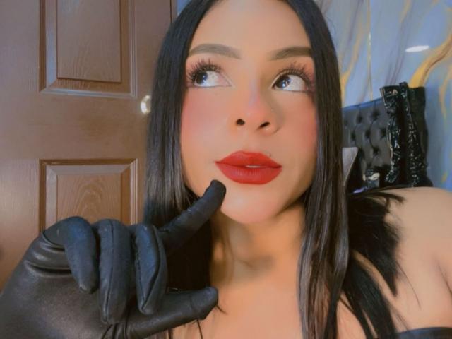 Nahaufnahme von LindaFrancoo mit schwarzen Haaren, rotem Lippenstift und schwarzen Handschuhen, wobei ein Finger ihre Lippe berührt und sie nach oben blickt. verführerischer Ausdruck, sexuelle Spannung, auffälliges Make-up, verführerischer Blick.