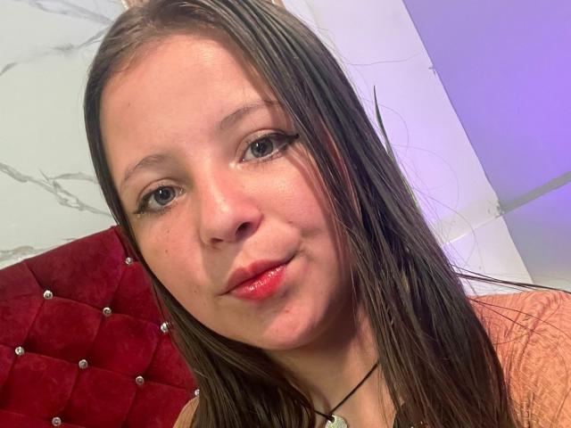 Close-up selfie of GraceRedMiller, light-skinned with straight brown hair, red lips, and blue eyes, leaning slightly forward. Red tufted headboard in the background, bright white wall on the right.