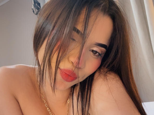 Close-up of LunaVali, a sexy woman with long brown hair, red lipstick, and heavy eyeliner, leaning forward with a suggestive expression. Her bare shoulder and gold necklace are visible. Fucking hot.