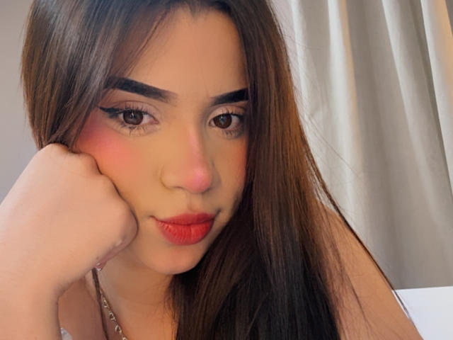 Gros plan sur LunaVali, une jeune femme aux longs cheveux bruns, au rouge à lèvres rouge et au trait d'eye-liner noir prononcé, qui pose sa joue sur sa main. Son expression est sensuelle et légèrement espiègle..