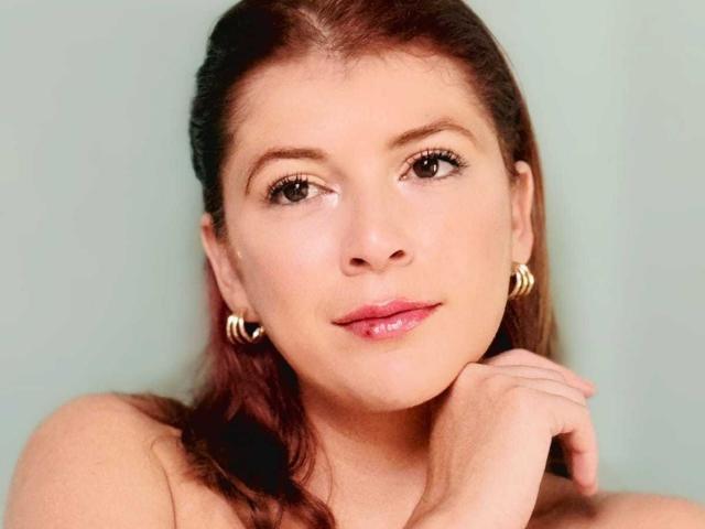 Close-up photograph of MoonXVelvet with brown hair, fair skin, and subtle makeup. She rests her chin on her hand, looking slightly to the side with a subtle smile. Wearing hoop earrings. Sexual tension.