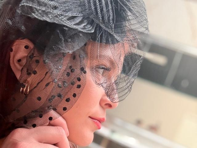 Close-up photo of MoonXVelvet wearing a black mesh veil with polka dots, gazing sideways with a slight smile. Her red lips contrast with fair skin, evoking a sensual, alluring vibe.