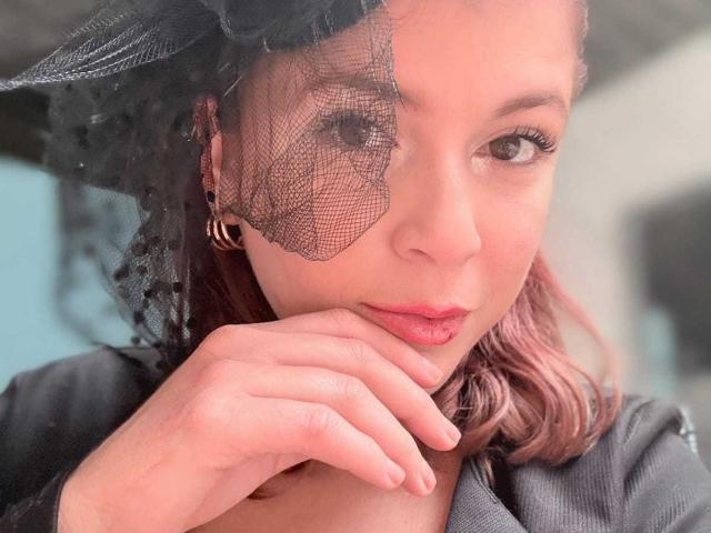 Close-up of MoonXVelvet with fair skin and pinkish-brown hair, wearing a black mesh eye mask, hand near chin, black clothing, intense gaze, slight smirk, soft light, sensual atmosphere.