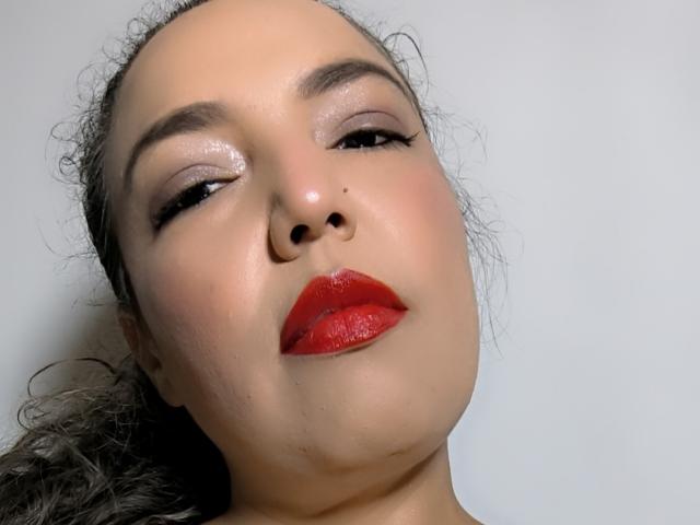 Close-up photo of Mariehss, a sexy woman with glossy, shiny skin and bright red lipstick, looking up with half-lidded eyes and a subtle smirk. Her dark hair is pulled back.