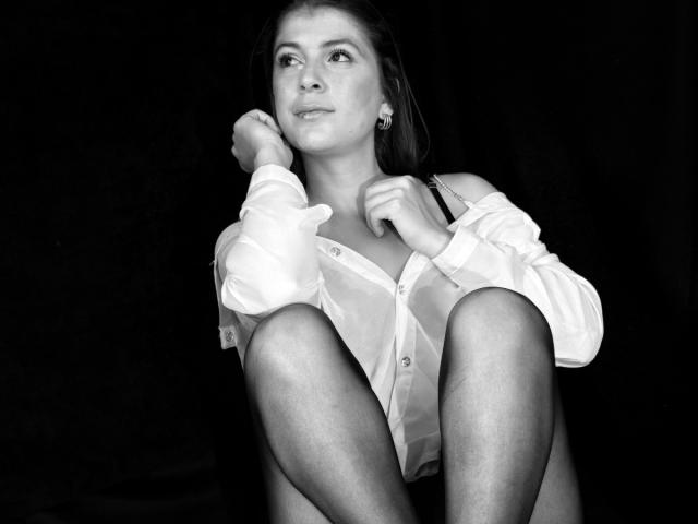 Schwarz-weißes Foto von MoonXVelvet, die mit angewinkelten Beinen sitzt und ein weißes Hemd trägt, das aufgeknöpft ist und so das Dekolleté freilegt. Hände in der Nähe des Gesichts, intensiver Blick, hoher Kontrast, sexy, verführerisch, provokative Pose..