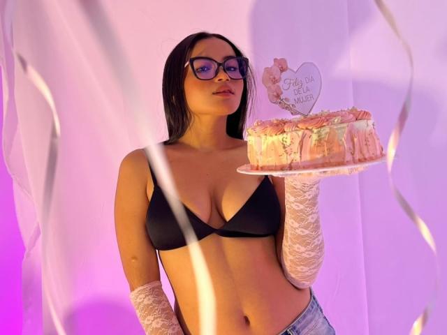 MiiaPalmer, aux cheveux foncés, portant un soutien-gorge noir et des gants en dentelle blanche, tient un gâteau éclairé en rose avec un petit décor en forme de cœur portant l'inscription "Just for U". Fond violet néon et rose. Gâteau en premier plan, aspect séduisant et ciblé..